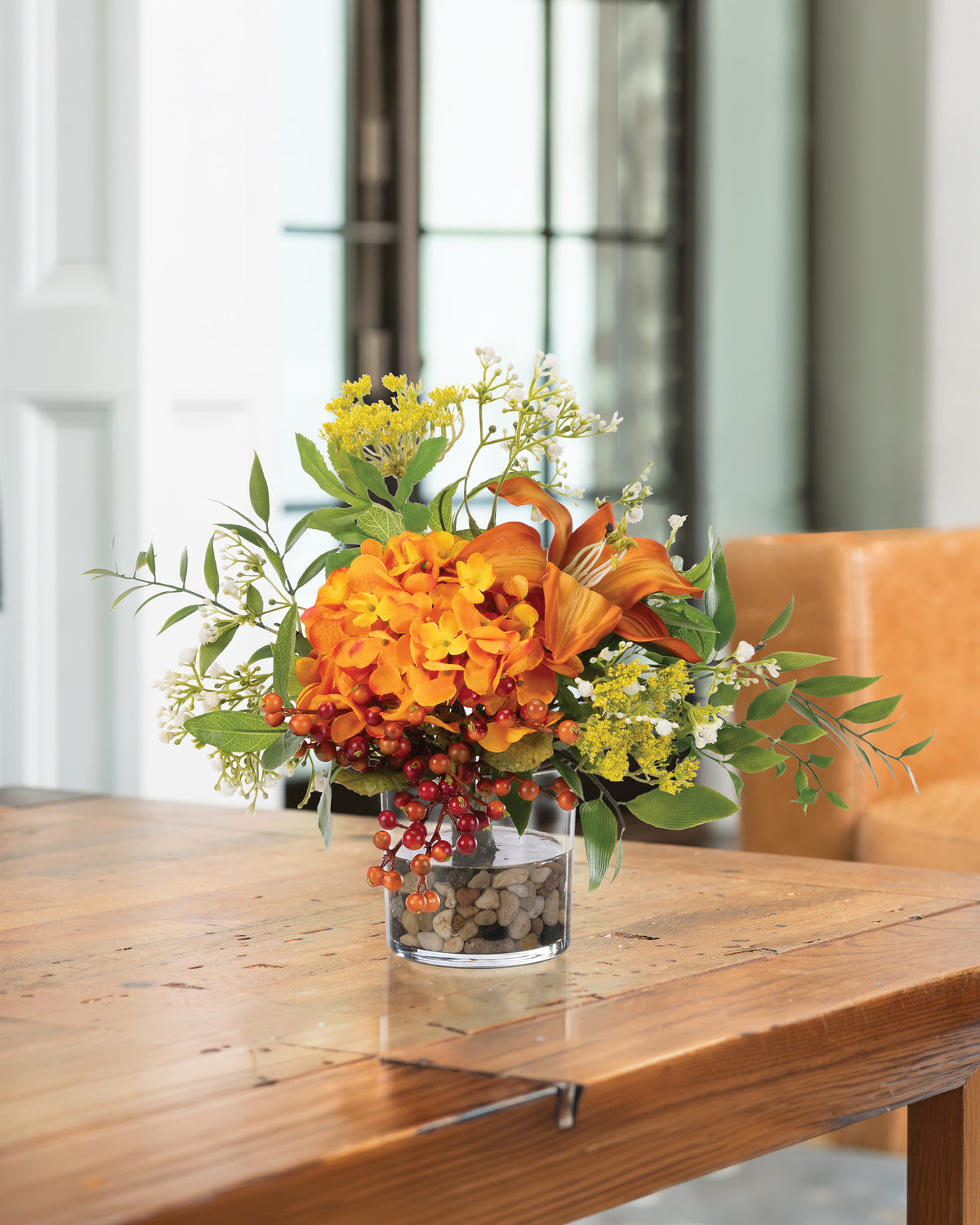 Hydrangea, Lilies & Berries Faux Flower Arrangement in a 4” glass vase of clear acrylic water and river stones displayed on a wooden table.