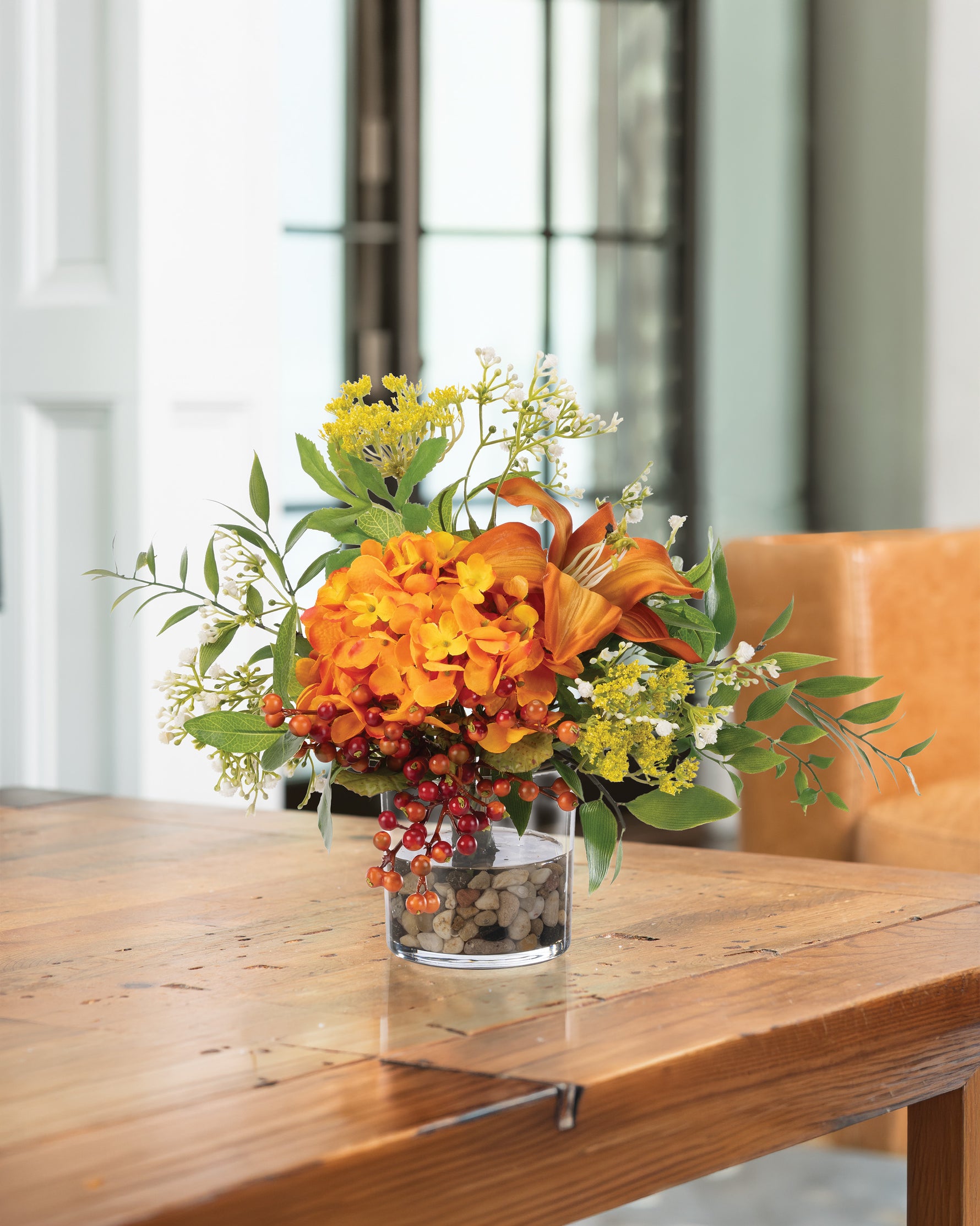 Hydrangea, Lilies & Berries Faux Flower Arrangement in a 4” glass vase of clear acrylic water and river stones displayed on a wooden table.