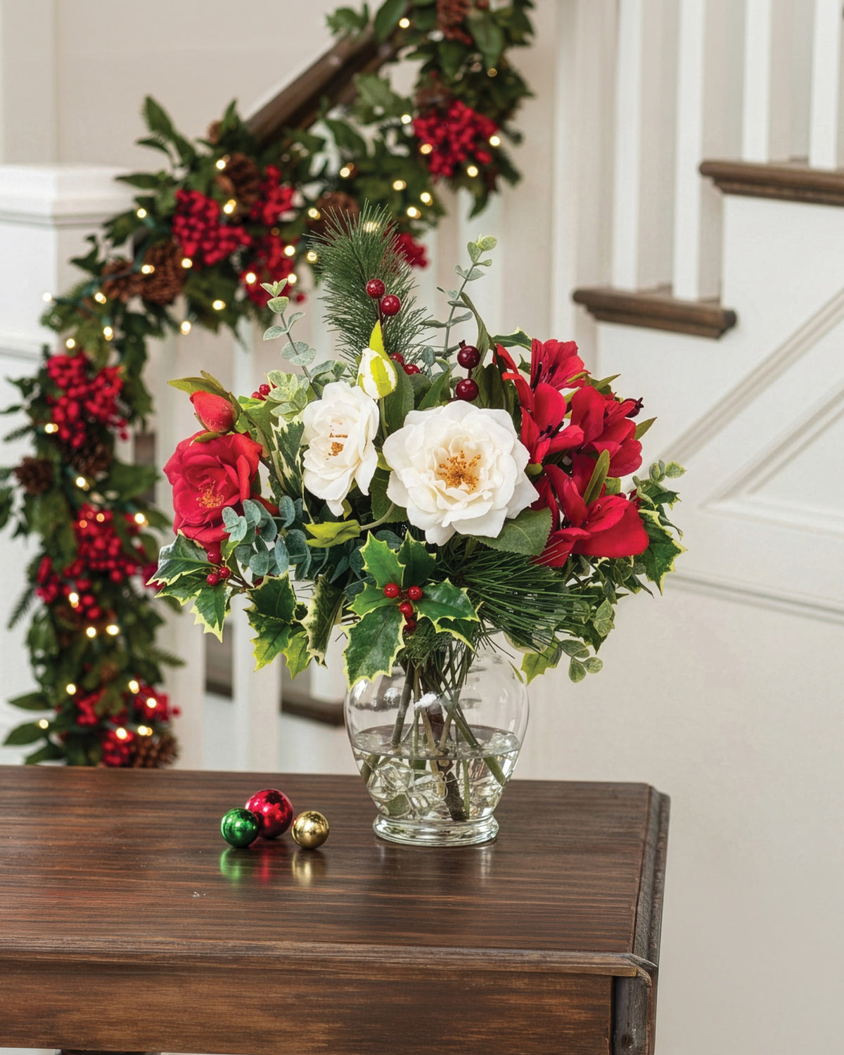 Holly, Pine & Berries Artificial Holiday Accent, set in a Glass Ginger Vase on a wooden table with christmas wreath on staircase.