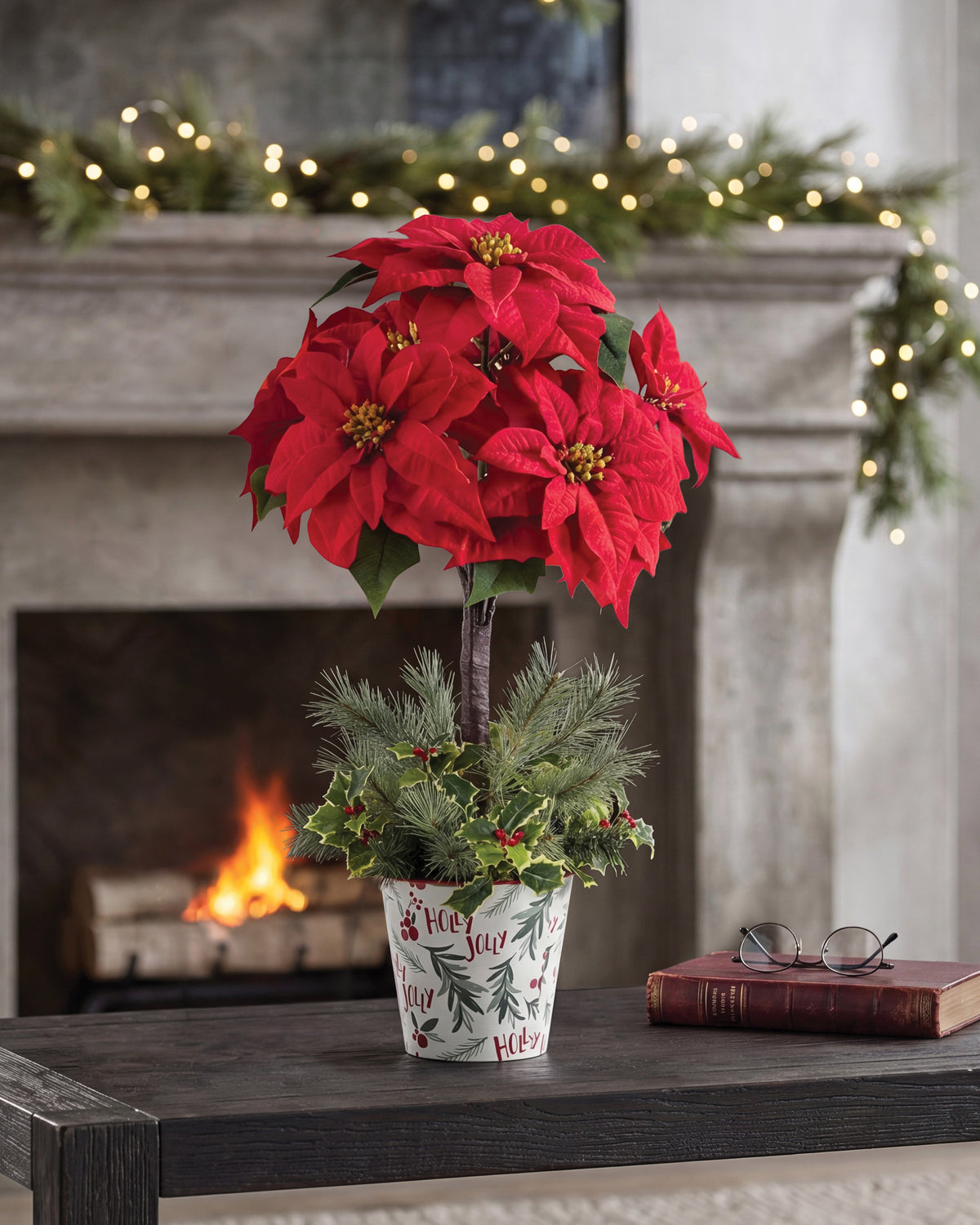 Silk Poinsettia Topiary, displayed in a 7” metal retro pot with lush evergreens, variegated holly, and berries on wooden table and surrounding by Christmas Decorations. Closeup