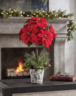 Silk Poinsettia Topiary, displayed in a 7” metal retro pot with lush evergreens, variegated holly, and berries on wooden table and surrounding by Christmas Decorations. Closeup