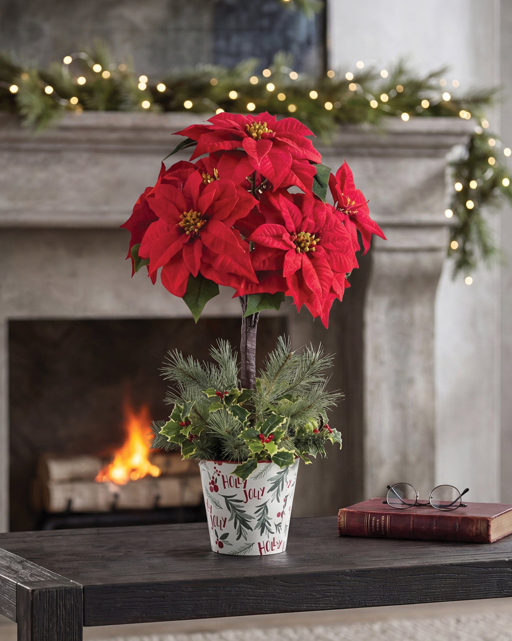 Silk Poinsettia Topiary, displayed in a 7” metal retro pot with lush evergreens, variegated holly, and berries on wooden table and surrounding by Christmas Decorations. Closeup