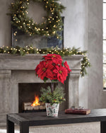 Silk Poinsettia Topiary, displayed in a 7” metal retro pot with lush evergreens, variegated holly, and berries on wooden table and surrounding by Christmas Decorations. 