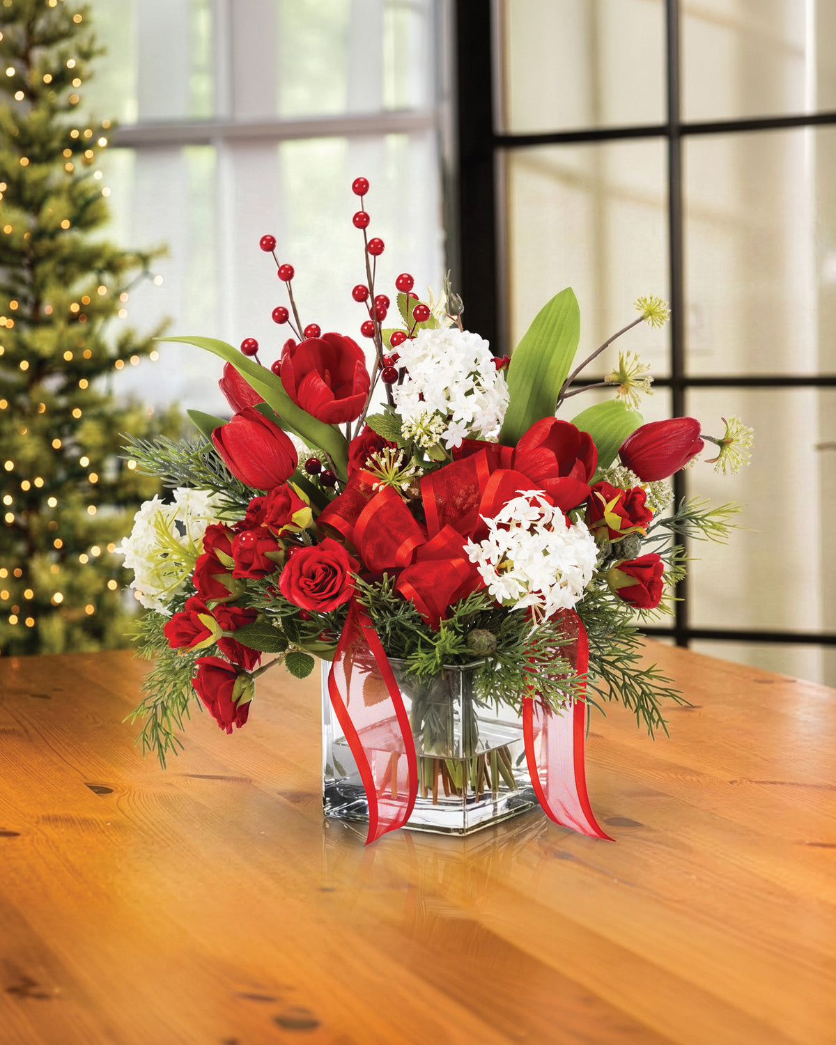 Tulip, Rose & Snowball Silk Flower Holiday Arrangement, nestled in a 4” square glass vase of clear acrylic water on a wooden table and Christmas Tree in the Background.