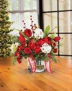 Tulip, Rose & Snowball Silk Flower Holiday Arrangement, nestled in a 4” square glass vase of clear acrylic water on a wooden table and Christmas Tree in the Background.