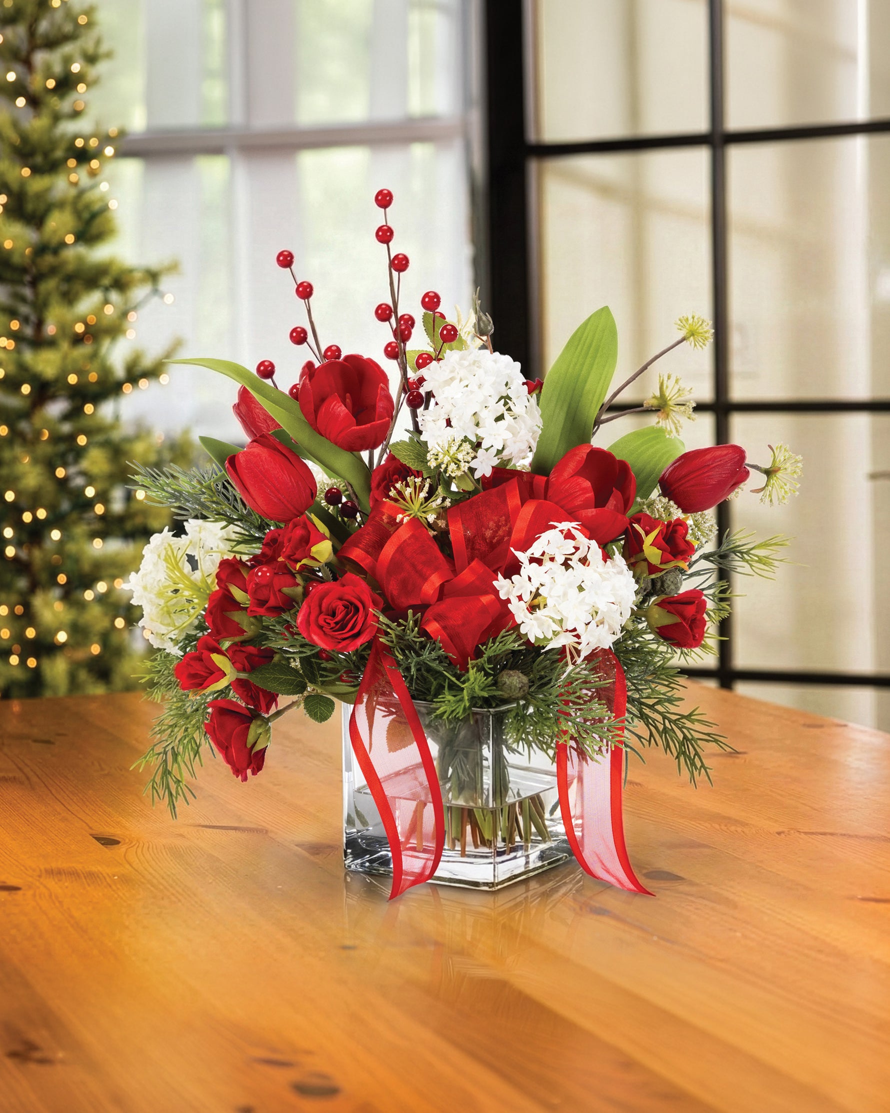 Tulip, Rose & Snowball Silk Flower Holiday Arrangement, nestled in a 4” square glass vase of clear acrylic water on a wooden table and Christmas Tree in the Background.