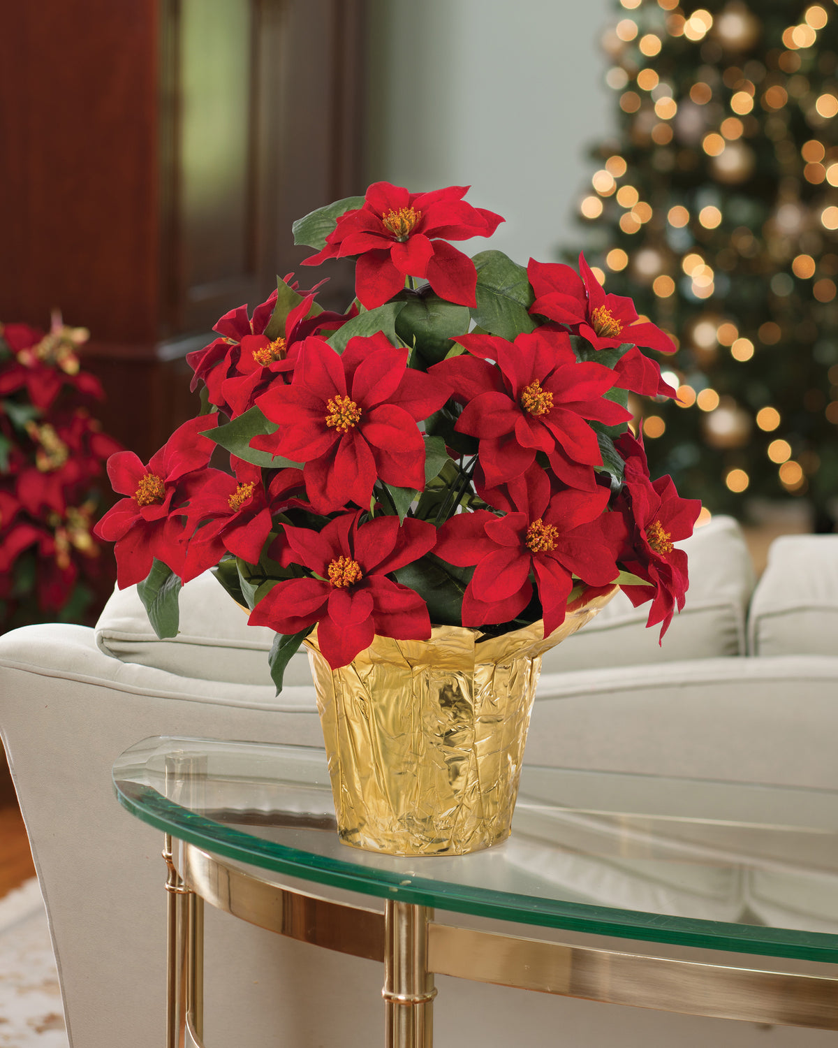 Desk Top Silk Poinsettia, potted in 4" Weighted Flower Pot with Shiny Gold Cover Included, set on a glass table and Christmas Decorations in the background. Closeup.
